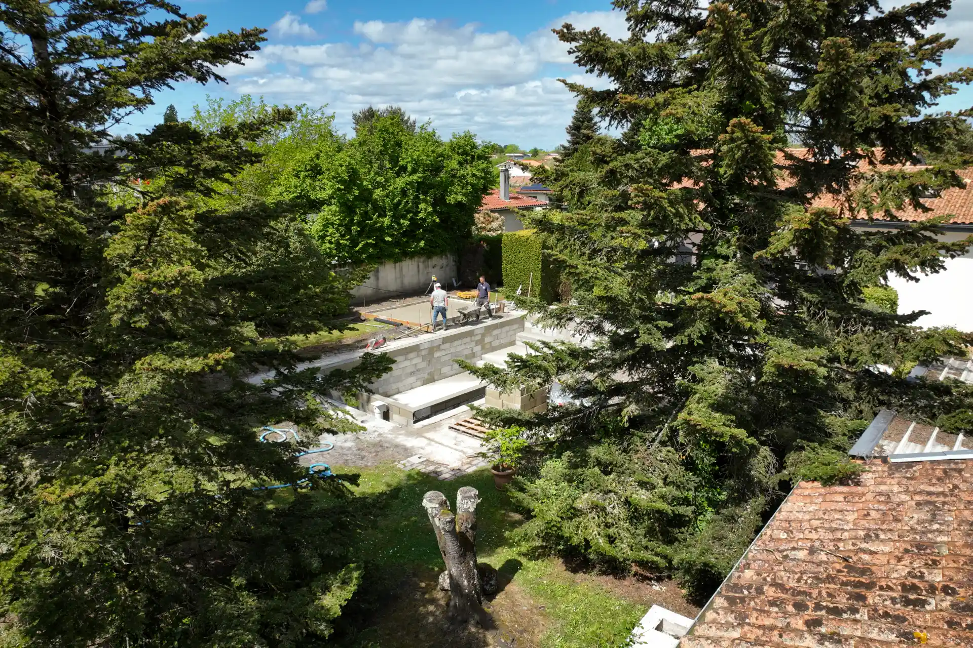 Vue aérienne discrète, à travers les arbres, d’ouvriers montant les murs en parpaings d’une piscine en construction